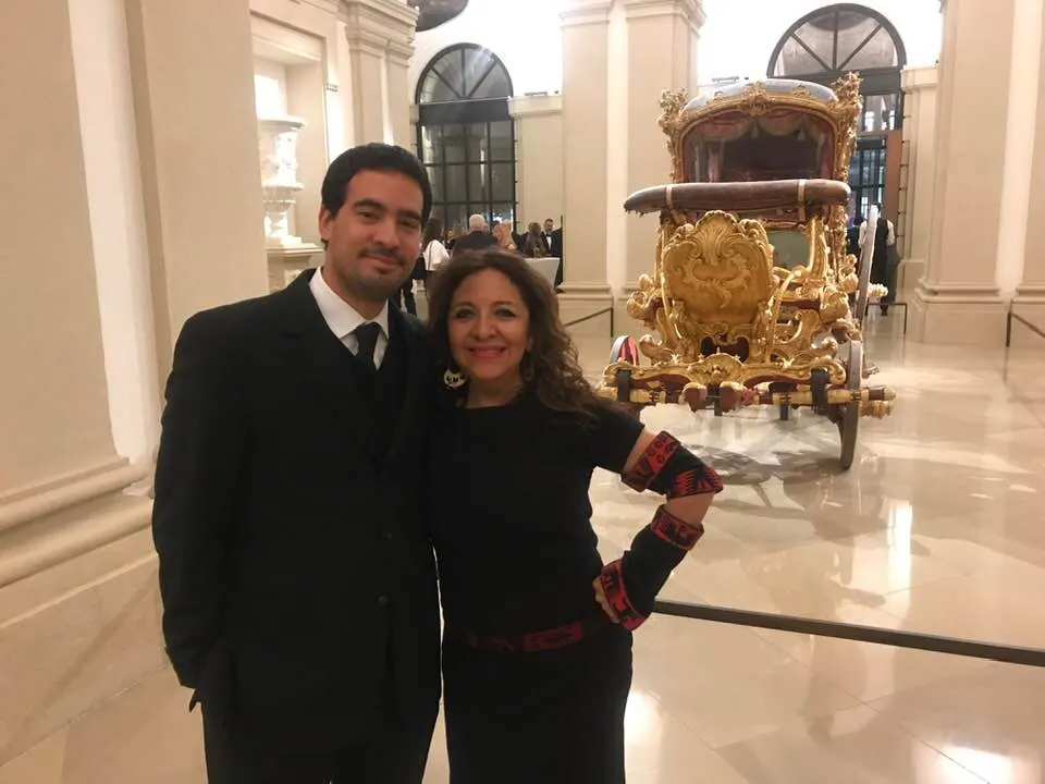 Dos asistentes frente a un carruaje dorado en un salón histórico
